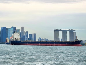 A large cargo ship is sailing on a body of water with a cityscape in the background, featuring modern skyscrapers and a distinctive three-towered hotel with a rooftop terrace.