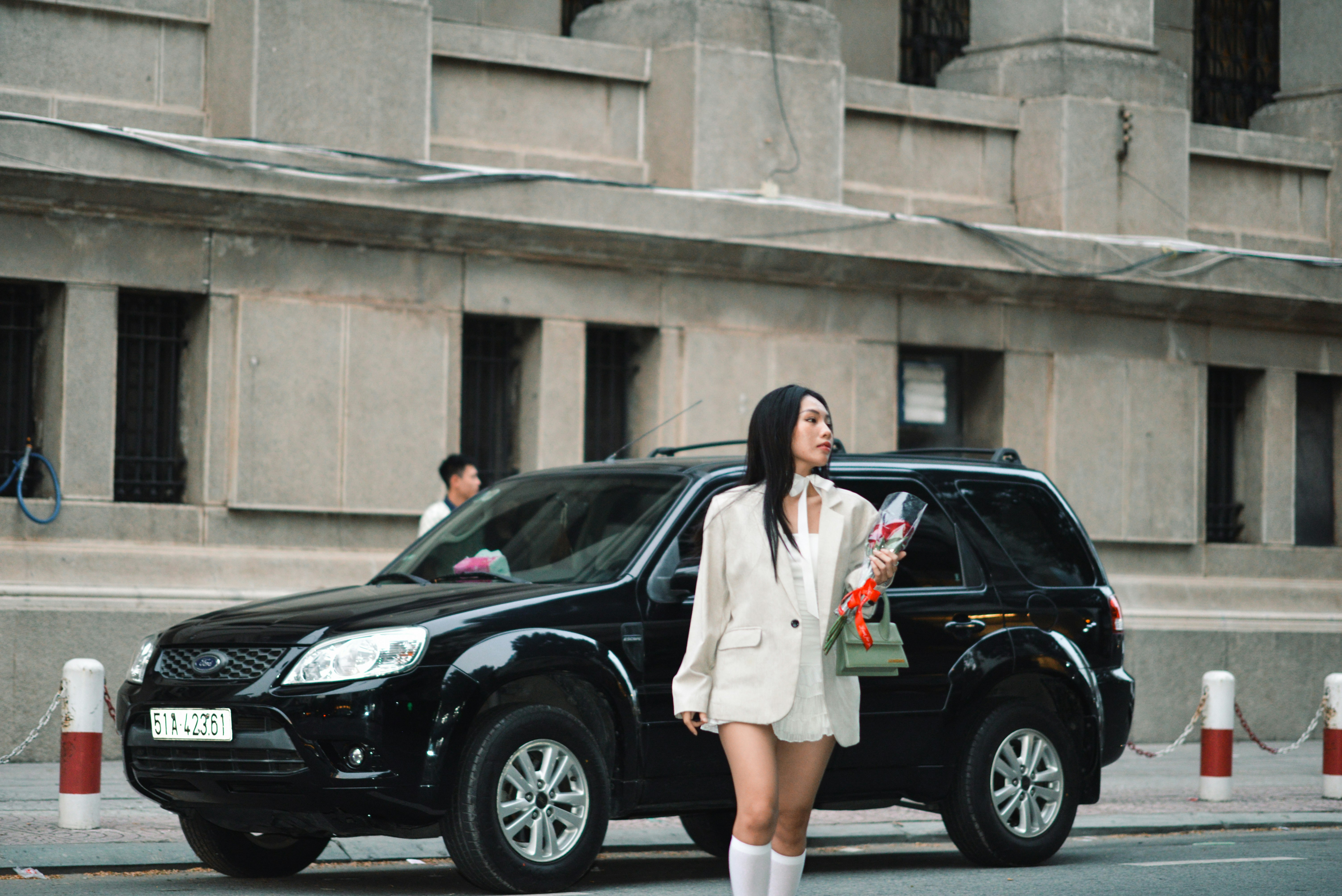 A woman in a white blazer and knee-high socks stands on the street holding a bouquet of red roses, next to a parked black SUV. There are large concrete buildings in the background.