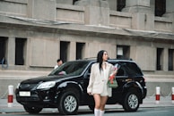 A woman in a white blazer and knee-high socks stands on the street holding a bouquet of red roses, next to a parked black SUV. There are large concrete buildings in the background.