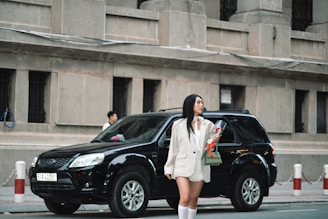 A woman in a white blazer and knee-high socks stands on the street holding a bouquet of red roses, next to a parked black SUV. There are large concrete buildings in the background.