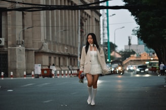 A woman in a white dress with a beige blazer and white knee-high socks walks down a city street in the evening. The background features a large building with columns on the left and a row of cars driving on the road. The lighting suggests an urban environment with streetlights reflecting off the wet pavement.