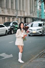 A woman wearing a light-colored blazer and shorts, accompanied by knee-high socks and pointed shoes, is standing by the roadside holding a bouquet of flowers. Cars are visible in the background, with an urban building lining the street. She appears to be smiling.
