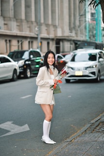 A woman wearing a light-colored blazer and shorts, accompanied by knee-high socks and pointed shoes, is standing by the roadside holding a bouquet of flowers. Cars are visible in the background, with an urban building lining the street. She appears to be smiling.