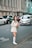 A woman wearing a light-colored blazer and shorts, accompanied by knee-high socks and pointed shoes, is standing by the roadside holding a bouquet of flowers. Cars are visible in the background, with an urban building lining the street. She appears to be smiling.