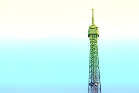 A stylized depiction of the Eiffel Tower dominates the frame, rising against a pastel gradient sky. The structure is infused with shades of green and blue, contrasting with the soft yellow and cyan sky, creating a surreal and artistic effect.