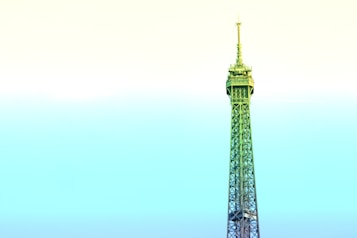 A stylized depiction of the Eiffel Tower dominates the frame, rising against a pastel gradient sky. The structure is infused with shades of green and blue, contrasting with the soft yellow and cyan sky, creating a surreal and artistic effect.