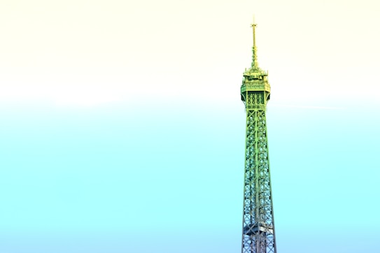A stylized depiction of the Eiffel Tower dominates the frame, rising against a pastel gradient sky. The structure is infused with shades of green and blue, contrasting with the soft yellow and cyan sky, creating a surreal and artistic effect.