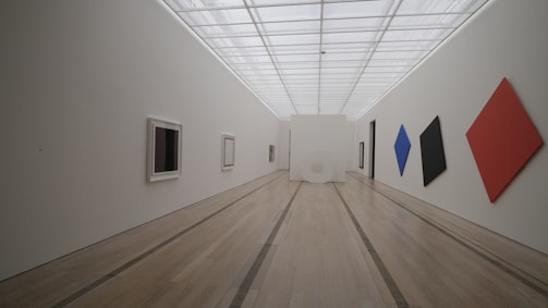A spacious, minimalist gallery features a high ceiling with a grid-like pattern and a wooden floor. The walls are adorned with modern art pieces, including diamond-shaped canvases in red, blue, and black, along with square and rectangular frames. The ambiance is clean and quiet, emphasizing the geometric artwork.