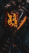 A dark-toned image featuring a motorcycle with a stylized, prominent seat design that includes the text 'Street Culture' in bold, yellow lettering. The words are surrounded by an orange geometric pattern, providing a vibrant contrast against the black background. Part of the motorcycle's handlebar and frame are visible, adding to the urban aesthetic.
