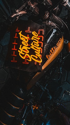 A dark-toned image featuring a motorcycle with a stylized, prominent seat design that includes the text 'Street Culture' in bold, yellow lettering. The words are surrounded by an orange geometric pattern, providing a vibrant contrast against the black background. Part of the motorcycle's handlebar and frame are visible, adding to the urban aesthetic.