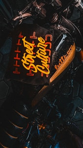 A dark-toned image featuring a motorcycle with a stylized, prominent seat design that includes the text 'Street Culture' in bold, yellow lettering. The words are surrounded by an orange geometric pattern, providing a vibrant contrast against the black background. Part of the motorcycle's handlebar and frame are visible, adding to the urban aesthetic.