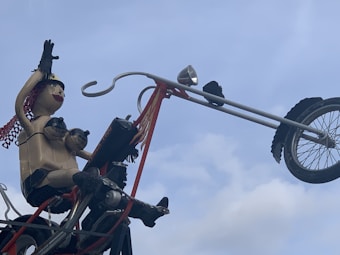 A sculpture of a person made from various materials is sitting on a long motorcycle handlebar. The figure has exaggerated features, including protruding eyes and breasts, and its arms are raised as if waving. The sculpture is set against a cloudy sky, giving it a whimsical appearance.