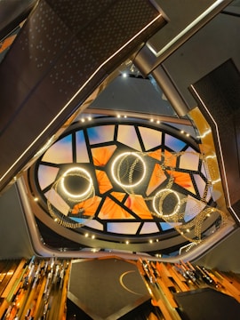 The image captures an abstract architectural interior with a modern and artistic ceiling design. The ceiling features an array of illuminated circular and semi-circular patterns, complemented by a mosaic of triangular segments. Below the ceiling, there are metallic structural frames and possible light fixtures adding to the contemporary aesthetic.