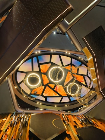 The image captures an abstract architectural interior with a modern and artistic ceiling design. The ceiling features an array of illuminated circular and semi-circular patterns, complemented by a mosaic of triangular segments. Below the ceiling, there are metallic structural frames and possible light fixtures adding to the contemporary aesthetic.
