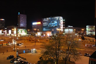A bustling cityscape at night featuring illuminated billboards prominently displaying advertisements for cars. The scene includes several multi-story buildings surrounded by illuminated streetlights and light trails from moving vehicles. Trees and parked cars are visible in the foreground, adding depth to the urban environment.