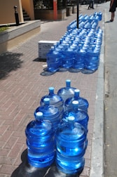 A large number of blue water jugs are lined up neatly on a sidewalk. The jugs reflect sunlight, creating a bright and shiny appearance. There are also a couple of white cardboard boxes among the jugs. The setting appears to be a sunny day in an outdoor urban environment with a paved walkway and curb visible.