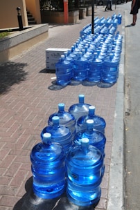 A large number of blue water jugs are lined up neatly on a sidewalk. The jugs reflect sunlight, creating a bright and shiny appearance. There are also a couple of white cardboard boxes among the jugs. The setting appears to be a sunny day in an outdoor urban environment with a paved walkway and curb visible.