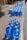 A large number of blue water jugs are lined up neatly on a sidewalk. The jugs reflect sunlight, creating a bright and shiny appearance. There are also a couple of white cardboard boxes among the jugs. The setting appears to be a sunny day in an outdoor urban environment with a paved walkway and curb visible.