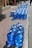 A large number of blue water jugs are lined up neatly on a sidewalk. The jugs reflect sunlight, creating a bright and shiny appearance. There are also a couple of white cardboard boxes among the jugs. The setting appears to be a sunny day in an outdoor urban environment with a paved walkway and curb visible.