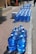 A large number of blue water jugs are lined up neatly on a sidewalk. The jugs reflect sunlight, creating a bright and shiny appearance. There are also a couple of white cardboard boxes among the jugs. The setting appears to be a sunny day in an outdoor urban environment with a paved walkway and curb visible.