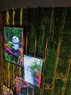 Several illuminated frames on a lush green vertical garden display vibrant images of a panda holding bamboo and a giraffe near a waterfall. The wall is composed of dense greenery, giving a feeling of a jungle environment.