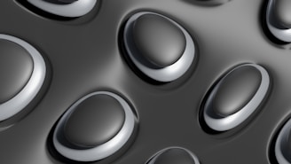 A close-up pattern of convex, oval-shaped structures with smooth surfaces on a dark, glossy background. The elliptical forms have a metallic appearance and are arranged in a repetitive, uniform manner.