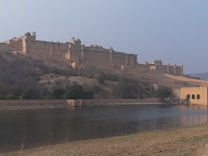 A large, historic fort complex with grand architectural features is situated on a hillside. It overlooks a body of water with a reflection of the fort visible. The landscape is arid, featuring sparse vegetation around the fort.