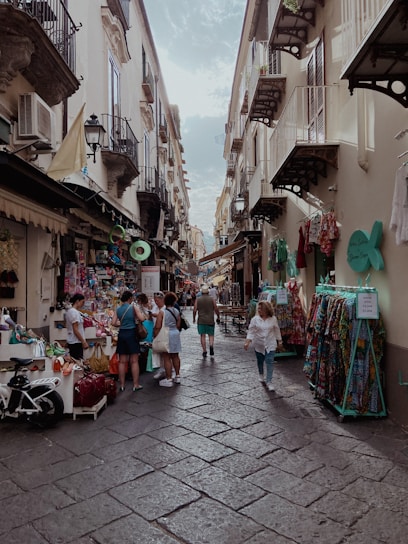 A narrow street lined with shops offering colorful clothing and other goods. People are casually walking and browsing around the street, which is bordered by old buildings with balconies. The sky is partially cloudy, and the atmosphere is lively and bustling.