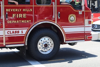 A red fire truck with the text 'Beverly Hills Fire Department' and 'Class 1' displayed on the side. The vehicle's large tires are visible, and there is a person inside the truck with their hand resting on the window frame.