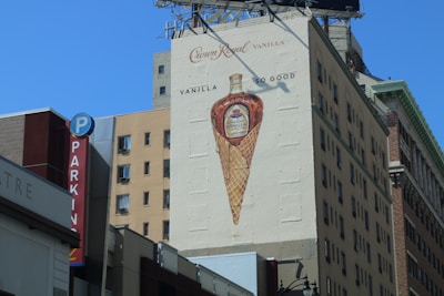 A large outdoor advertisement is painted on the side of a building, depicting a whiskey bottle inside a waffle cone. The ad promotes Crown Royal Vanilla, with the tagline 'Vanilla So Good.' Surrounding the mural are various buildings, including one with a vertical parking sign.