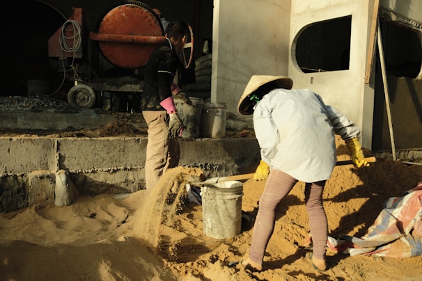 Two people are working on a construction site, moving sand with shovels and using buckets. One person is wearing a conical hat and the other is partially visible, with machinery and building structures in the background.