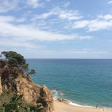 A tranquil ocean view with clear blue waters meeting the horizon, surrounded by a partly cloudy sky. In the foreground, a rocky cliff adorned with greenery juts into the scene. Below, a sandy beach stretches out with a few scattered people enjoying the coastline.