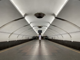 A modern underground subway platform characterized by its sleek, arched ceilings and symmetrical lighting. The platform is empty, creating a sense of spaciousness and stillness. Overhead lights are uniformly distributed along the ceiling, illuminating the clean and glossy tiled floor. Information panels and digital screens are visible above the tracks, contributing to the functional design of the station.