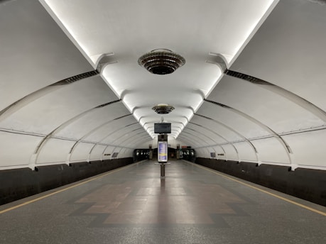 A modern underground subway platform characterized by its sleek, arched ceilings and symmetrical lighting. The platform is empty, creating a sense of spaciousness and stillness. Overhead lights are uniformly distributed along the ceiling, illuminating the clean and glossy tiled floor. Information panels and digital screens are visible above the tracks, contributing to the functional design of the station.