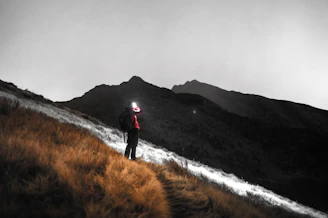 A rugged hiker using a high-lumen searchlight to illuminate a vast mountain trail at dusk