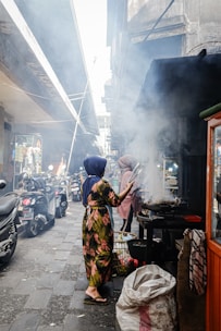 A bustling street scene with a woman cooking food at a street stall, surrounded by smoke from the grill. The woman is wearing a floral dress and a headscarf. Other people and motorbikes are visible along the narrow alleyway, which is lined with various shops and stands.