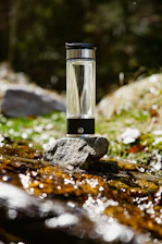 A bright pink-labeled Rosafonte water bottle resting on smooth stones by a clear stream.