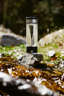 A transparent water bottle with a black cap is placed on a stone by a stream. The background features greenery and blurred foliage, suggesting a natural outdoor setting. Sunlight glistens off the water and the bottle, creating a reflective effect.