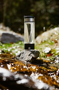 A transparent water bottle with a black cap is placed on a stone by a stream. The background features greenery and blurred foliage, suggesting a natural outdoor setting. Sunlight glistens off the water and the bottle, creating a reflective effect.