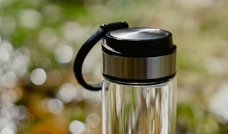 A clear glass water bottle with a stainless steel lid and black handle, set against a softly blurred background of green and white bokeh.
