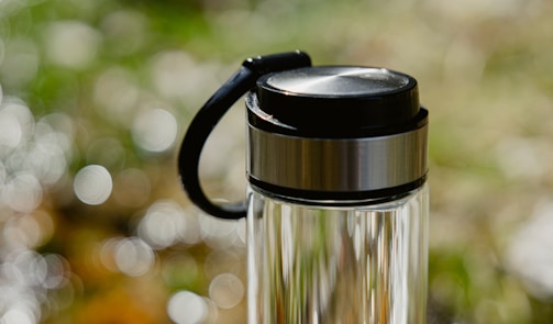 A clear glass water bottle with a stainless steel lid and black handle, set against a softly blurred background of green and white bokeh.
