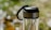 A clear glass water bottle with a stainless steel lid and black handle, set against a softly blurred background of green and white bokeh.