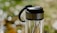 A clear glass water bottle with a stainless steel lid and black handle, set against a softly blurred background of green and white bokeh.