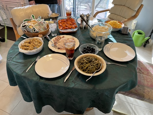 A round dining table covered with a dark green tablecloth holds a variety of dishes typically associated with a hearty meal. Dishes include sliced roast turkey, stuffing, green beans, mashed potatoes, sweet potatoes, black olives, corn, and rolls. There are plates and utensils set for a meal, along with glasses of iced tea. The background features chairs and kitchen cabinetry.