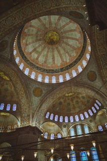 A grand, intricately adorned dome with elaborate geometric patterns and Arabic calligraphy. The dome is supported by ornate arches and columns, with stained glass windows letting in soft, colorful light. The architecture reflects traditional Islamic artistry, featuring detailed floral motifs and extensive decorative elements.