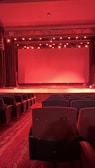 A dimly lit theater with rows of empty red seats facing a stage. The stage lighting casts a red hue across the entire scene, with numerous spotlights above shining downwards. Thick curtains flank the stage, and the flooring is carpeted with a patterned design.