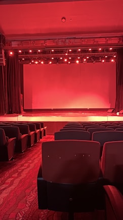 A dimly lit theater with rows of empty red seats facing a stage. The stage lighting casts a red hue across the entire scene, with numerous spotlights above shining downwards. Thick curtains flank the stage, and the flooring is carpeted with a patterned design.