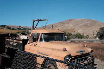 A vintage tow truck with a weathered orange paint job is parked behind a chain-link fence. The truck is marked with 'Dons Antique Auto Parts' and rests in a setting with dry grass and rolling hills under a clear blue sky. The hills in the background have the word 'NILES' marked on one of them.