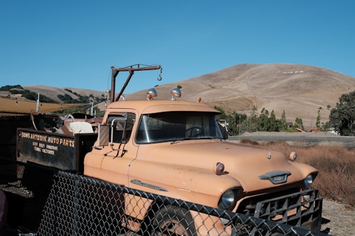A vintage tow truck with a weathered orange paint job is parked behind a chain-link fence. The truck is marked with 'Dons Antique Auto Parts' and rests in a setting with dry grass and rolling hills under a clear blue sky. The hills in the background have the word 'NILES' marked on one of them.
