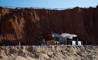 A small beachside café with a dark wooden exterior and white umbrellas is nestled against a towering cliff of reddish-brown rock. Sun loungers and a red parasol are visible on the sandy area in front, while the café has a small elevated terrace with railing.